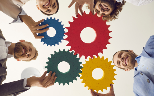 Four people smiling and holding colorful gears, viewed from below, symbolizing teamwork and collaboration.