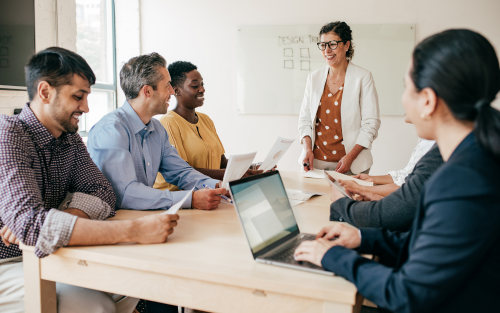 A diverse group of professionals having a meeting around a table, with one person standing and presenting.