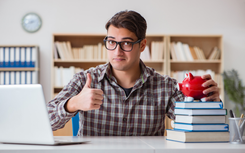 Here's the short alt text for the image you provided:  "Man in glasses giving a thumbs-up while holding a piggy bank and sitting at a desk with books and a laptop."