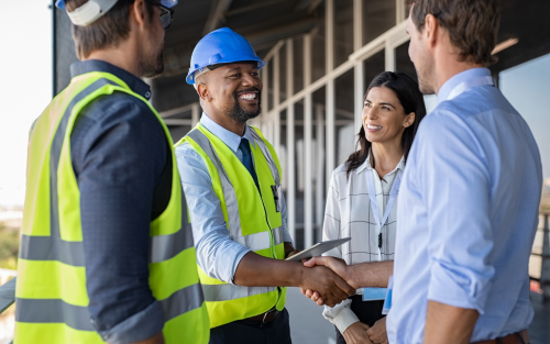 Construction workers in safety gear shaking hands with business professionals at a construction site.