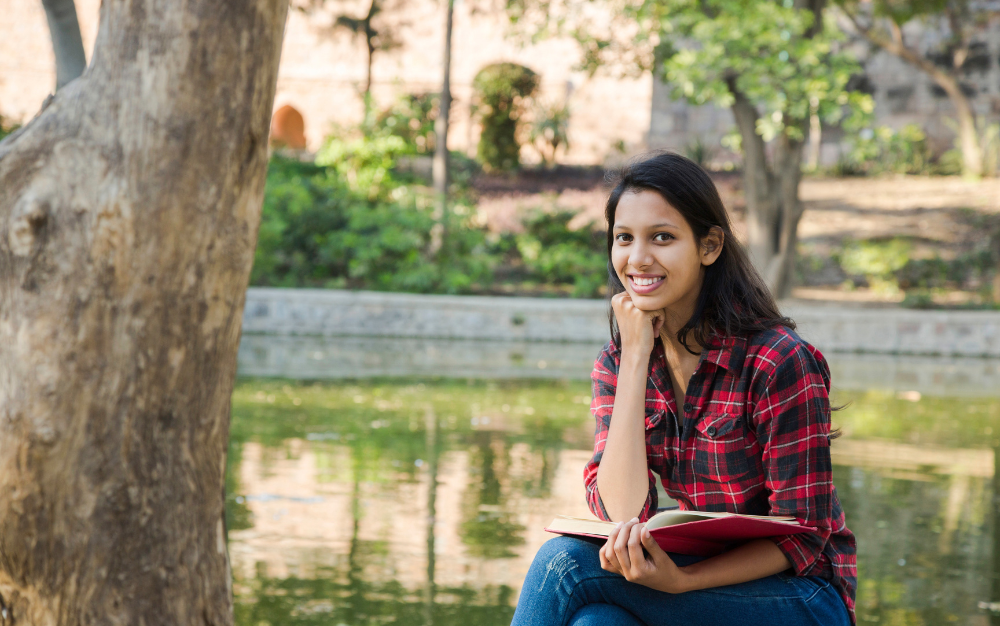 Here's the short alt text for the image you provided:  "Smiling woman in a red plaid shirt holding an open book outdoors near a pond."