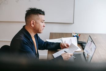 A man sits at a desk, focused on his laptop while holding a piece of paper. He appears to be working in a professional setting.